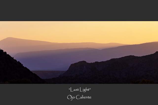 A shadowed landscape photograph of Ojo Caliente, the silhouette of the mountains fading into soft sunlight