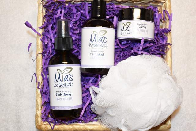 Three different sized black containers with white labels sit in a wicker bin atop purple decorative packaging, next to a white loofa. Labeling reads "Mias Botanicals Aromatherapy, Sheer Essence 2 in 1 wash"