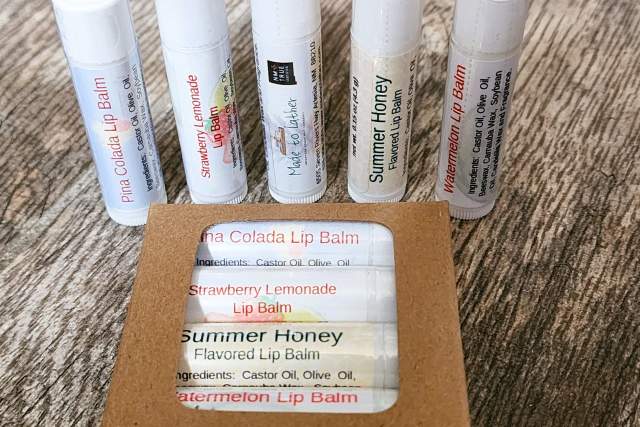 9 different flavors of lip-balm in white tubes with light coloring sit on a wooden table. Five tubes are standing upright behind another 4 within a minimalist cardboard package.