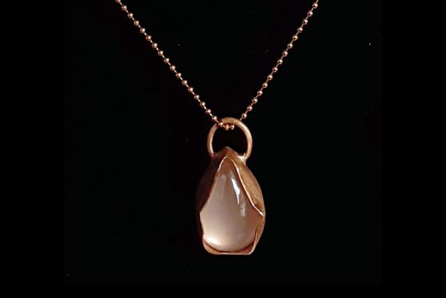 a copper strapped necklace encompasses a white gemstone, it hangs in front of a black background