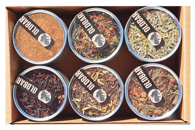 six silver tins with clear tops sit inside a cardboard box, containing various spices and herbs