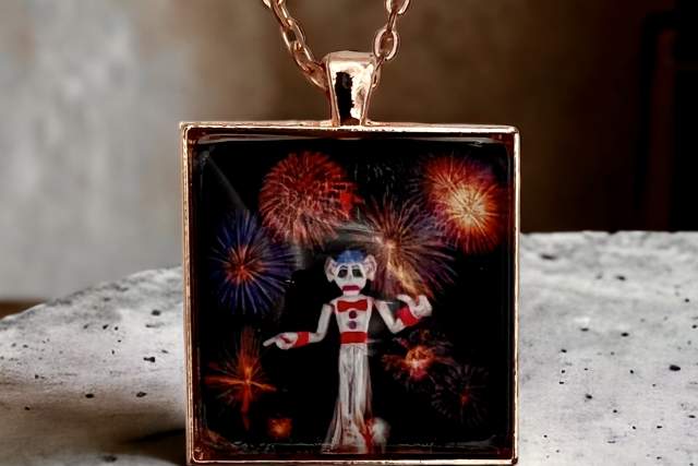 a pendant with a copper border and chain depicts the towering white effigy "zozobra" with fireworks bursting in the background. it sits on a white granite countertop