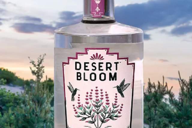 A bottle of "Desert Bloom Purple Sage" gin is displayed outdoors against a scenic background of a sunset sky and green foliage. The label features illustrations of sage and hops.