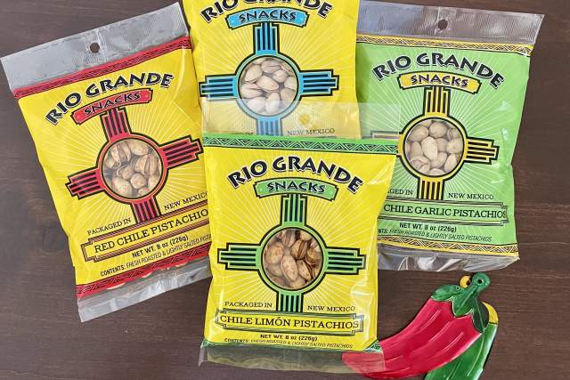 Three bags of Rio Grande Snacks pistachios in different flavors—Red Chile Pistachio, Naturally Raw Pistachio, and Chile Limón Pistachio—displayed on a wooden surface with a decorative chili pepper ornament beside them.