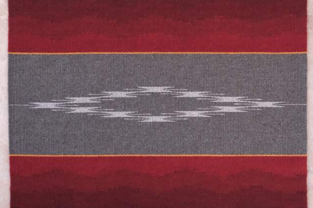 a red, grey and white navajo rug with a diamond design in the middle sits flat on a white surface