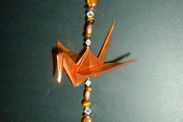 A copper origami crane is suspended by a string with orange and white beads. A small white tag is affixed at the top of the line