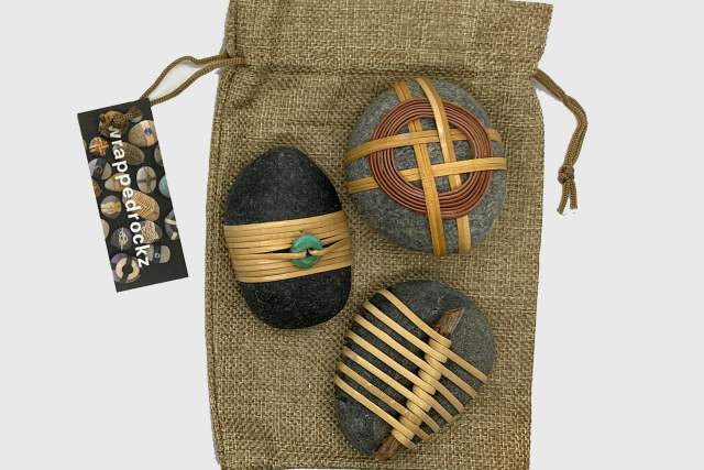 black and grey stones wrapped in twine, accentuated by sticks and turquoise sit on a brown back. The tag on the bag reads wrapped rockz