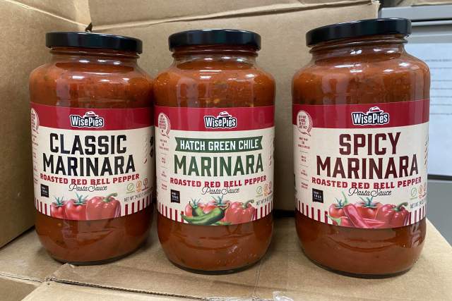 Three different bottles of marinara sauce sit in clear jars on a cardboard box. Labels read from left to right "Classic Marinara, Hatch Green Chile Marinara, Spicy Marinara"