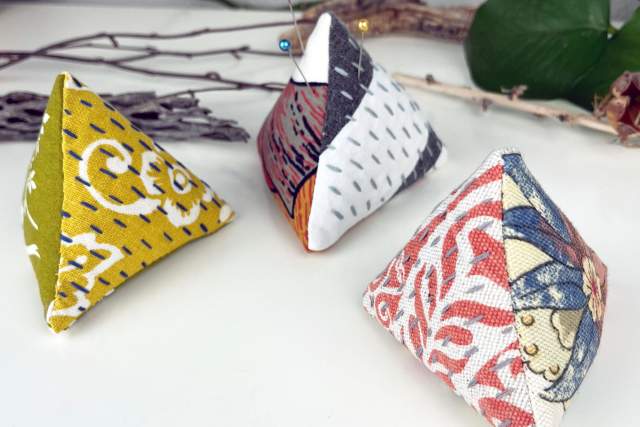 Colorful triangular pincushions with unique patterns sit on a white table with sticks and leaves in the background