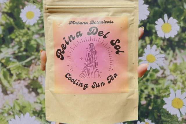 A brown bag with a pink label is being held in front of a mural of flowers. The label reads "Reina Del Sol, Cooling Sun Tea"