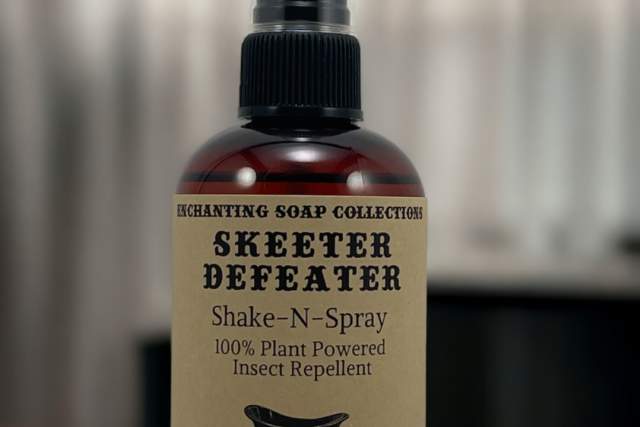 A brown spray bottle sits on a wooden desk in front of white linen curtains. The tan label on the bottle reads "Enchanting Soap Collection, Skeeter Defeater, Shake-N-Spray, 100% Plant Powered Insect Repellent"