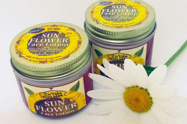 two multicolored containers sit next to a white flower on a white background. Label has a sunflower on it and reads "Sun Flower Face Lotion"