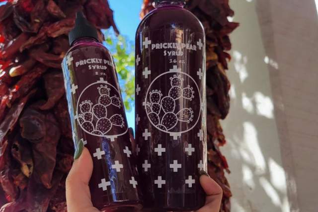Two clear bottles filled with red syrup are held in front of two Ristras hung on a white porch. Label reads "Prickly Pear Syrup"