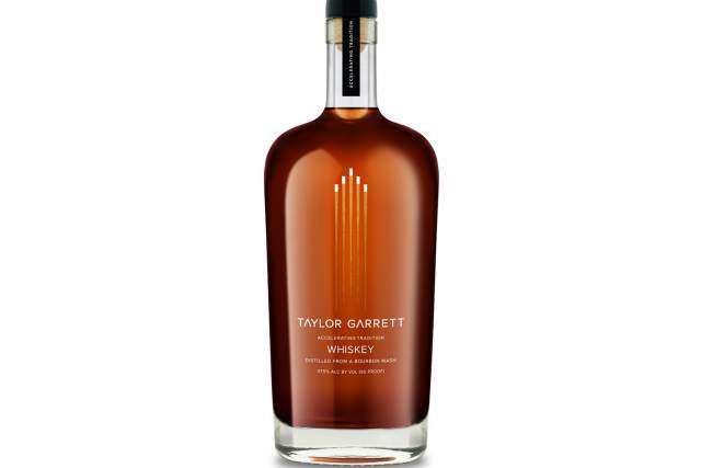 A clear bottle of whiskey, filled with brown whiskey. Text reads Taylor Garett Whiskey, Accelerating Tradition, Distilled from a bourbon mash