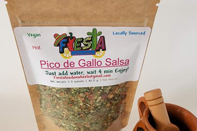 A brown and clear bag with dried salsa ingredients sits next to a brown mortar and pestle. The label reads "Fiesta Food On Wheels, Vegan, Hot, Locally Sourced, Pico de Gallo Salsa, Just add water and wait 4 mins"