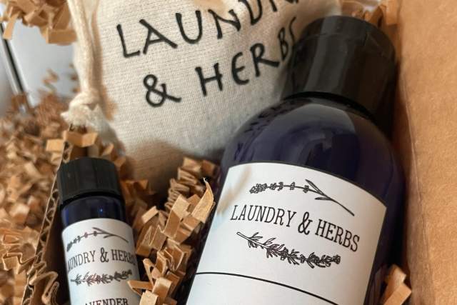 A bottle of "Laundry & Herbs" lavender laundry detergent is nestled in crinkled paper packaging. A small vial labeled "Lavender" and a drawstring bag with the "Laundry & Herbs" logo are nearby.