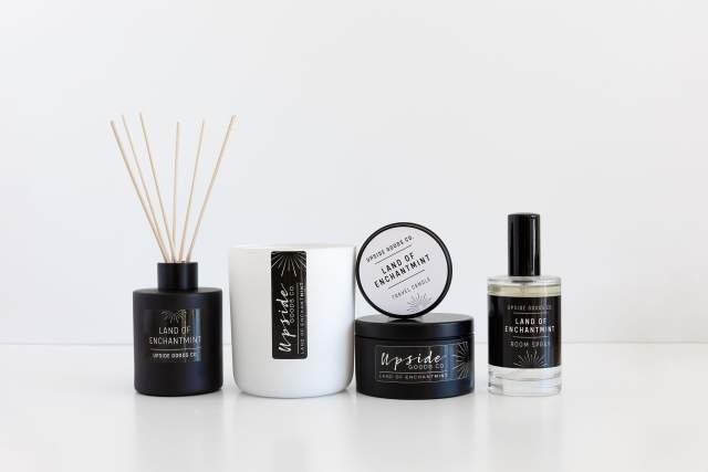 A black incense holder with wooden sticks, white candle with black label, black container and a clear spray bottle with black labeling. Text reads "Upside Goods Co. Land of Enchantmint"