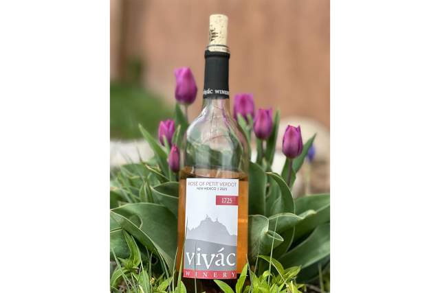 A clear bottle with pink wine 3/4 full, has a cork sticking out of the black neck of the bottle. The label is pink and white, and depicts a minimalist outline of a mountain. The Label reads Rose of Petit Verdot, New Mexico - 2023. 1725, Vivac Winery