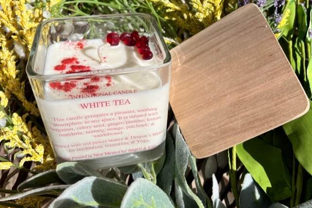 A clear container filled with white and red wax sits on a bed of green and yellow plants with a wooden lid next to it. Label reads "Intentional Candle, White Tea"