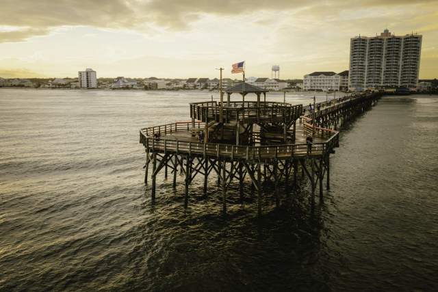 Cherry Grove Pier stretches out into the ocean at sunset in North Myrtle Beach, with an American flag flying above its distinctive octagonal deck and covered pavilion. A few people are seen enjoying the view and fishing along the wooden structure. The calm waves reflect the golden sky, while beachfront condos and homes line the shoreline in the background.
