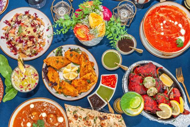 An overhead view shows a full table spread of multiple Indian dishes, including curry, naan bread, appetizers, sauces, and beverages.