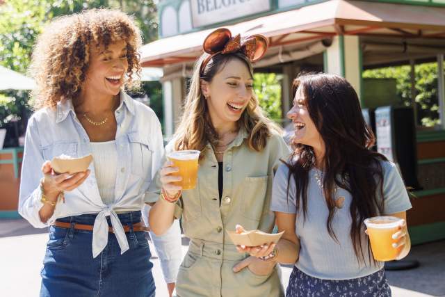 Three women walking at EPCOT enjoying drinks and food from a festival booth.