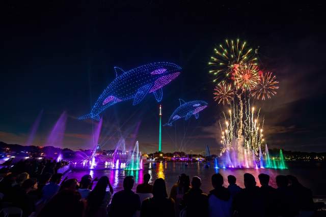 Drone lights form orca shapes above a lagoon during a nighttime show at SeaWorld Orlando, accompanied by fireworks and illuminated fountains. Guests are seated along the waterfront viewing area as multicolored lighting effects and water features enhance the performance.