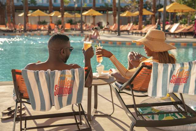 A man and a woman unwind by the pool at Universal Endless Summer Resort – Dockside Inn and Suites. They are seen from behind, raising celebratory cocktails while overlooking the sparkling pool.