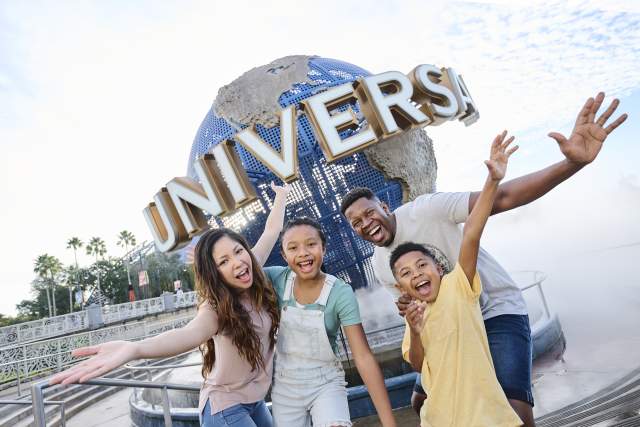 A smiling family of four — mom, dad, preteen daughter and preteen son — stand in front of the Universal globe, their arms outstretched and welcoming.