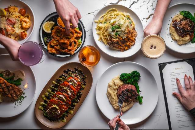 Selection of restaurant entrées, appetizers, pasta dishes, cocktails, and desserts arranged across a marble tabletop under decorative table lamps, showcasing a variety of menu offerings.