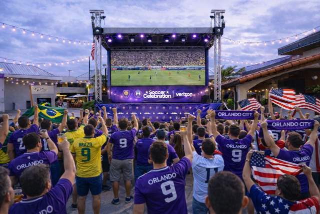 Renderings of viewing areas, Outdoor watch party with a crowd gathered in front of a large elevated screen displaying a soccer match. Attendees wear team jerseys and hold flags while standing beneath string lights and stage lighting in an open-air venue.