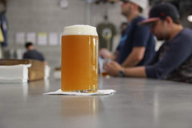Light, frothy beer in a glass, on countertop at the Crooked Can Brewing Company. Slightly off-center with people at the bar in the background.