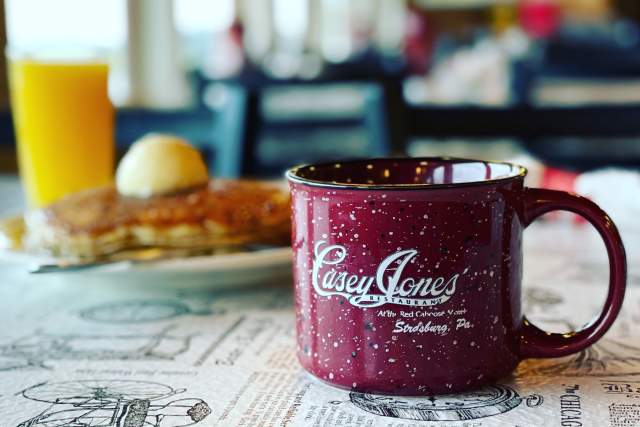 A red coffee mug that reads Casey Jones Restaurant with a glass or orange juice and a plate of pancakes with butter in the background