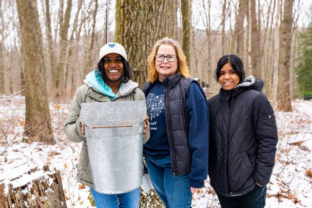 Three women holding up a bucket full of maple syrup in a wooded area with trees and snow on the ground