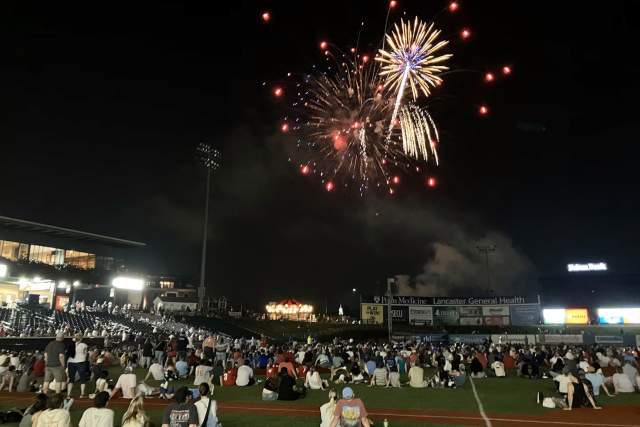 People sitting on a baseball field at night watching a firework display