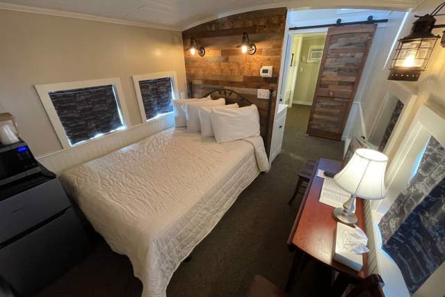Birds eye view of the interior of a motel bedroom with a full-sized bed, a dresser with a lamp, and a door to a bathroom.