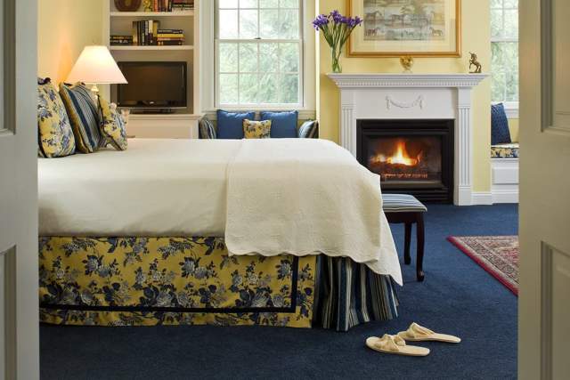 Interior of a bedroom at the Inn at Whitewing with a made bed, slippers on the ground, and a lit fireplace in the background