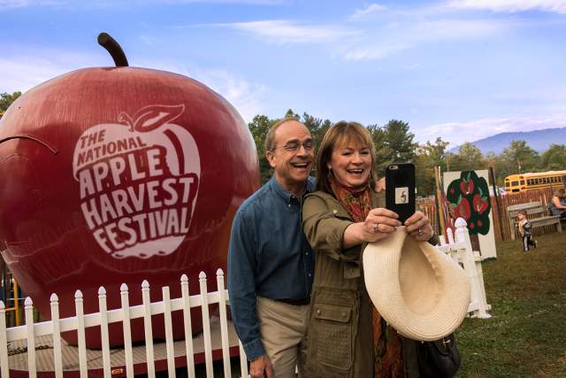 A man and woman taking a selfie with a giant plastic apple that reads The National Apple Harvest Festival