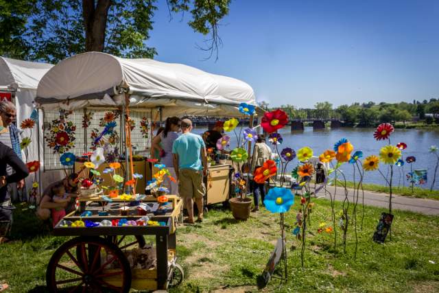 People shopping at a vendor cart with large, plastic flowers along a river