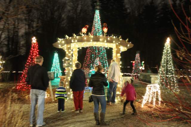 Family of six standing in front of a decorated archway with holiday lights and trees are lit with holiday lights behind the archway