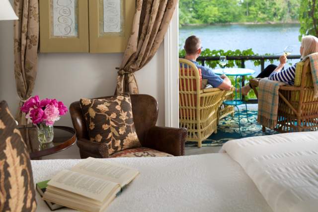 Book laid open on a bed with two people sitting in chairs on a balcony overlooking a river in the background.
