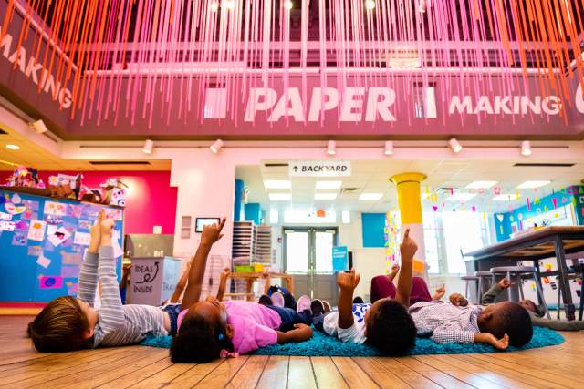 Kids lying on their backs in a classroom pointing up at the ceiling