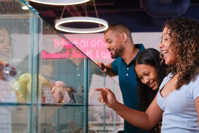 A girl, a woman, and a man looking and pointing and a glass case in an ice cream shop with different ice cream flavors
