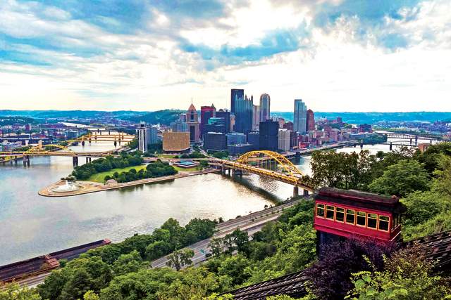 An aerial view of a red cable car moving down a side of a mountain with a skyline of Pittsburgh in the background