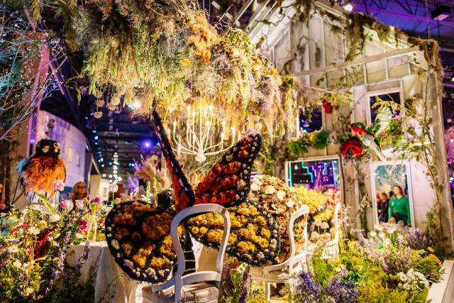A flower exhibit with a butterfly made of flowers sitting on a chair at a table with flowers hanging up above
