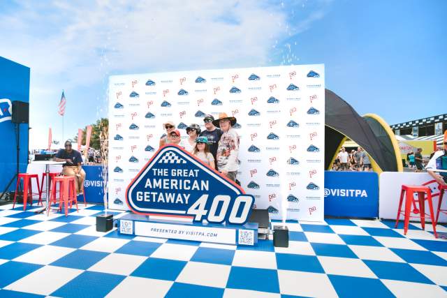 A family of five posing in front of a step and repeat with the Visit PA and Great American Getaway 400 logo while standing behind an enlarged statue of the Great American Getaway 400 logo.