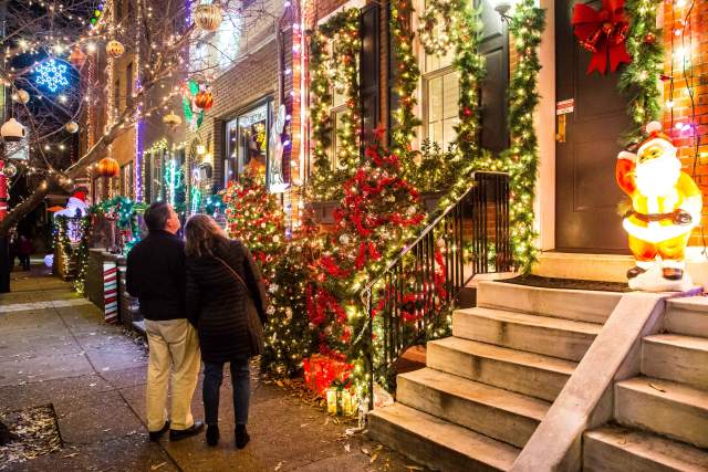 A man and woman holding hands on a sidewalk and looking up at townhouses with holiday lights.
