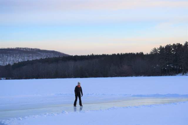 Girl in winter gear ice skating on a frozen pond with snow and a forest of trees in the background