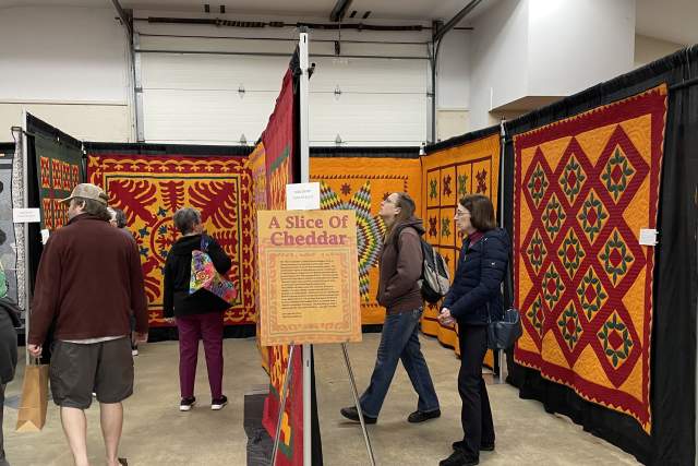People admiring quilts hung up on display at a quilt show. A sign in front of the display reads A Slice of Cheddar with a description about the quilts.