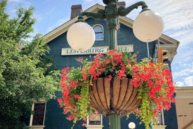 A lamp post with a potted flower attached to it in front of a quant blue house that reads Lewisburg Hotel